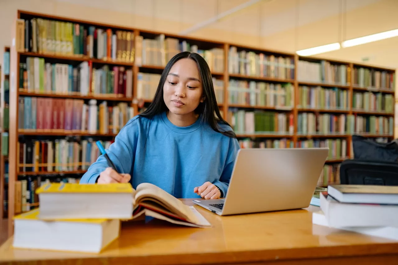 female college student sits at a desk in a library writing in a book